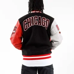 A man wearing a Men's Pro Standard NBA Chicago Bulls Varsity Jacket, showcasing a bold "CHICAGO" embroidery across the back with contrasting black, red, and gray elements.