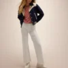 A model wearing the Mother The Western Varsity Jacket in navy blue with cream detailing, paired with white pants and a striped red shirt.
