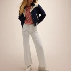 A model wearing the Mother The Western Varsity Jacket in navy blue with cream detailing, paired with white pants and a striped red shirt.