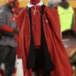 A football player wearing a long red cape-style coat with the Kansas City Chiefs logo on the sleeve, standing on the sidelines of a stadium.