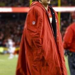 A football player bundled in a red cape-style coat, buttoned in the front, with a high collar and hood, standing in a cold-weather game setting.