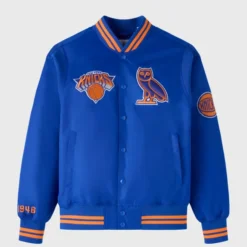OVO x NBA New York Knicks Satin Varsity Jacket front view, featuring Knicks and OVO owl patches on a blue satin body with orange detailing.