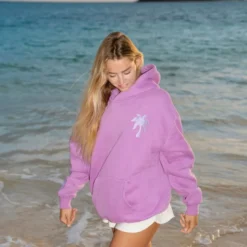 Woman wearing a pink "Pink Palm Puff Everything Comes in Waves Açai Hoodie" with a small palm tree logo on the chest, standing on a beach.