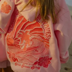 Pink hoodie featuring a bold red embroidered design of a surfer, palm tree, and waves with the phrase "Everything Comes in Waves.