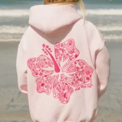 A woman standing on the beach wearing a pastel pink hoodie with a large pink hibiscus floral design on the back.