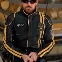 Ryan Gosling wearing a black NASA leather jacket with yellow stripes and space mission patches.